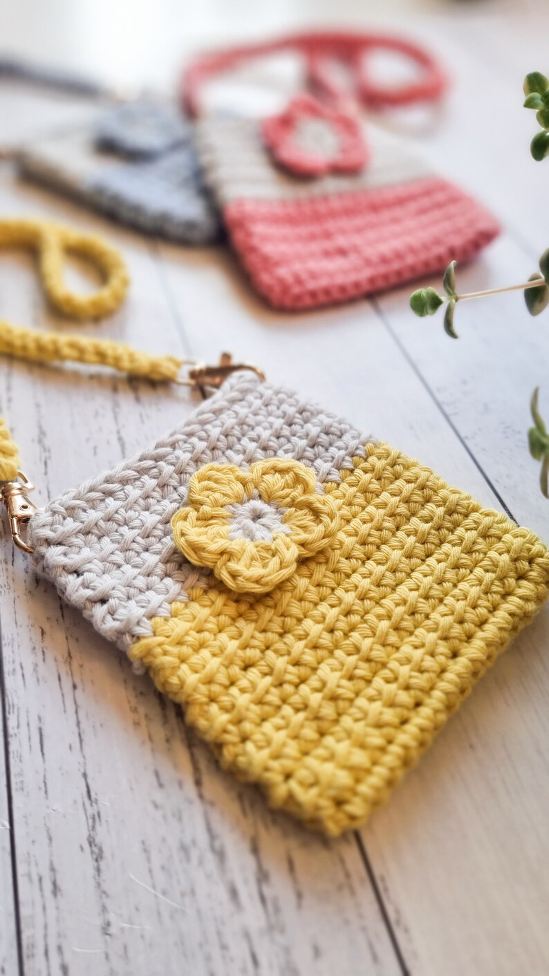 Pode incluir: Capa de telefone de croch&ecirc; feita &agrave; m&atilde;o em amarelo e bege com um detalhe floral amarelo. A capa tem uma al&ccedil;a de pulso e um fecho dourado. Outras capas de croch&ecirc; em rosa e azul s&atilde;o vis&iacute;veis no fundo.