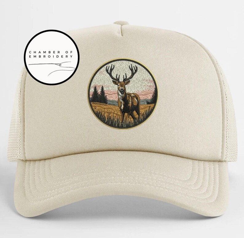 May include: A beige trucker hat featuring a circular embroidered patch on the front. The patch depicts a brown stag with large antlers standing in a field with trees and a sunset sky. The text CHAMBER OF EMBROIDERY is printed in a circle above.