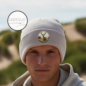 May include: A light beige knit beanie featuring a circular embroidered patch on the front. The patch depicts a stag standing in a grassy field with trees and a sunset sky in the background. The text Chamber of Embroidery is visible in a logo.