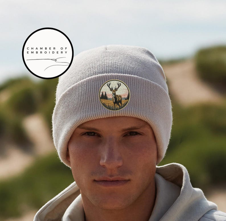 May include: A light beige knit beanie featuring a circular embroidered patch on the front. The patch depicts a stag standing in a grassy field with trees and a sunset sky in the background. The text Chamber of Embroidery is visible in a logo.