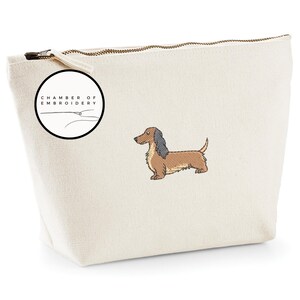 May include: Cream-colored canvas cosmetic bag with a brown and black dachshund embroidery. The bag has a zipper closure and a circular logo that says "Chamber of Embroidery". The dachshund is embroidered in detail.