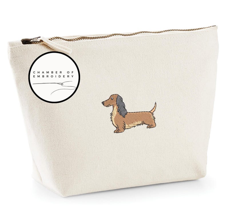 May include: Cream-colored canvas cosmetic bag with a brown and black dachshund embroidery. The bag has a zipper closure and a circular logo that says "Chamber of Embroidery". The dachshund is embroidered in detail.