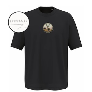 May include: A black short-sleeved t-shirt featuring a circular embroidered patch on the chest depicting a deer in a field with trees at sunset. A circular logo in the upper left corner contains the text CHAMBER OF EMBROIDERY with a needle icon.