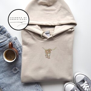 May include: A beige hoodie with a brown and white longhorn cow embroidered on the front. The hoodie has a drawstring hood and a kangaroo pocket. The text "Chamber of Embroidery" is on a white circle in the top left corner of the image.