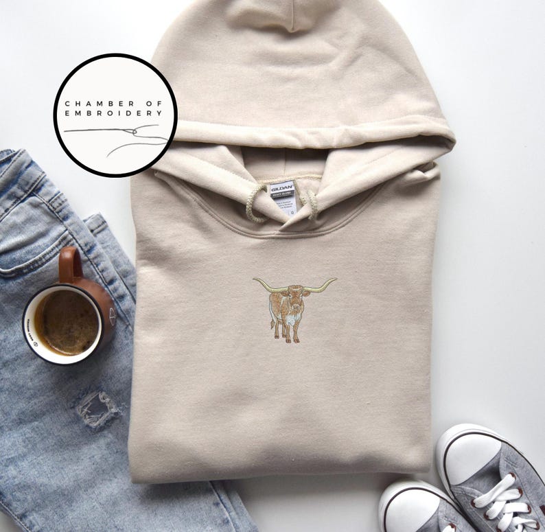 May include: A beige hoodie with a brown and white longhorn cow embroidered on the front. The hoodie has a drawstring hood and a kangaroo pocket. The text "Chamber of Embroidery" is on a white circle in the top left corner of the image.