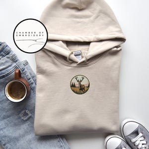 May include: A beige hooded sweatshirt featuring a circular embroidered patch on the chest. The patch depicts a deer standing in a grassy field with trees and a sunset sky. A logo circle with the text CHAMBER OF EMBROIDERY is visible above.