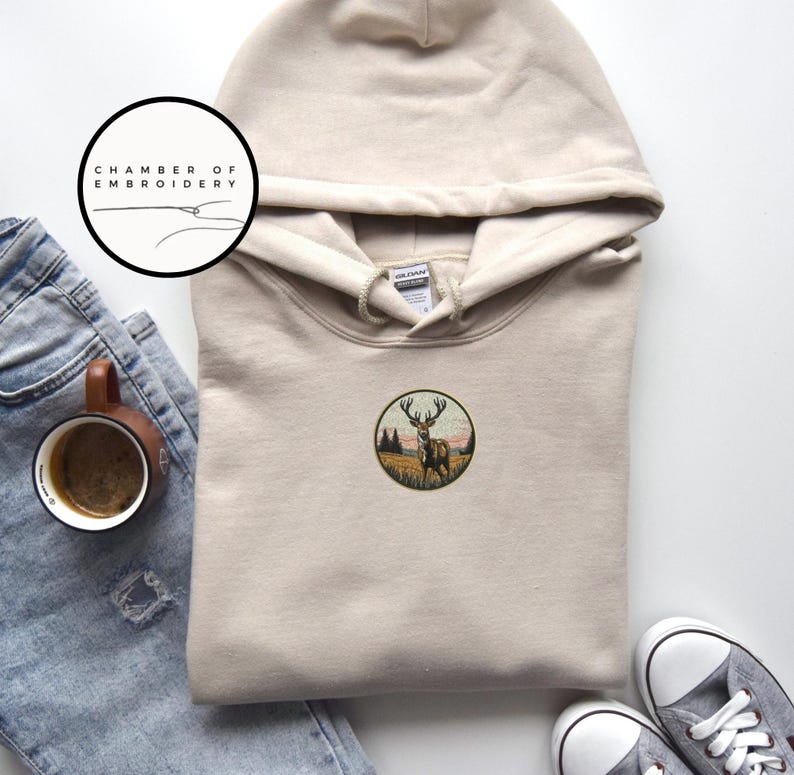 May include: A beige hooded sweatshirt featuring a circular embroidered patch on the chest. The patch depicts a deer standing in a grassy field with trees and a sunset sky. A logo circle with the text CHAMBER OF EMBROIDERY is visible above.