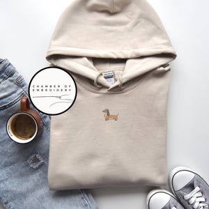 May include: A beige hooded sweatshirt with a small embroidered dachshund dog on the front. The sweatshirt has a drawstring hood and a kangaroo pocket. The image also includes a pair of gray sneakers, a cup of coffee, and a pair of blue jeans.