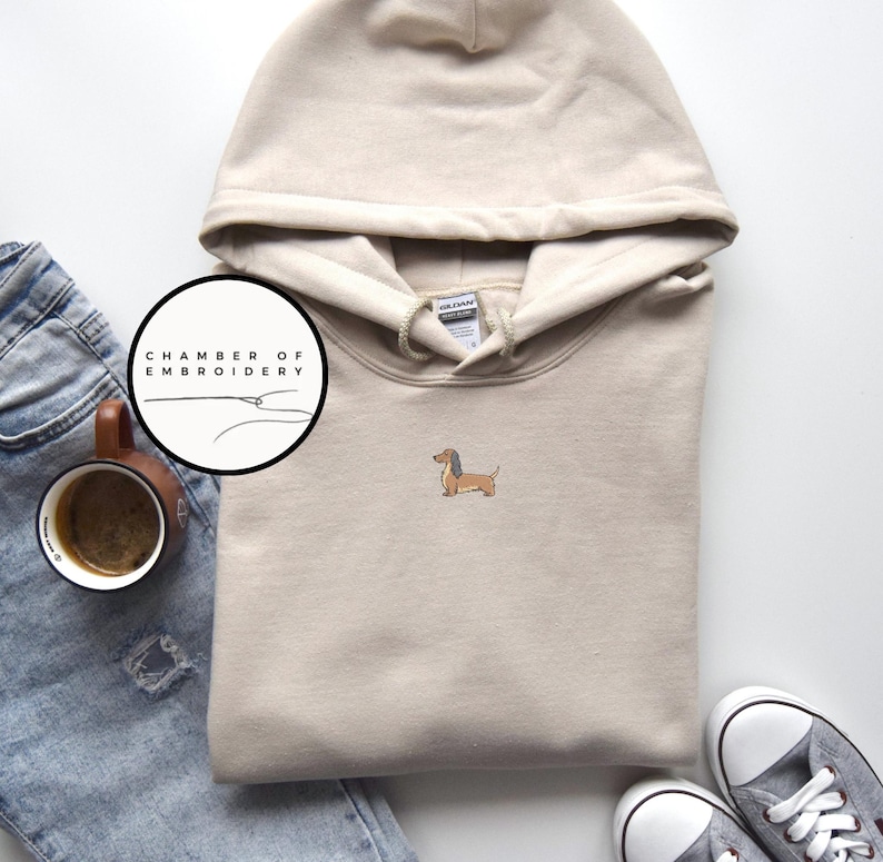 May include: A beige hooded sweatshirt with a small embroidered dachshund dog on the front. The sweatshirt has a drawstring hood and a kangaroo pocket. The image also includes a pair of gray sneakers, a cup of coffee, and a pair of blue jeans.
