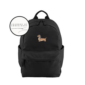 May include: Black backpack with a brown and tan dachshund dog embroidery. The backpack has a top handle, two shoulder straps, and a front pocket. A circular logo with the text "CHAMBER OF EMBROIDERY" is in the upper left corner.