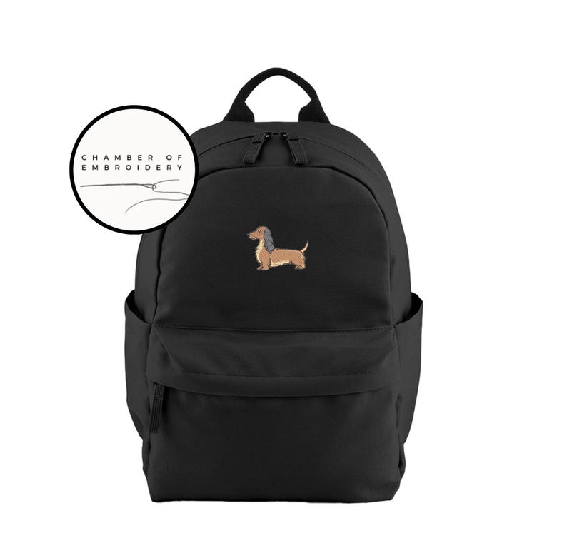 May include: Black backpack with a brown and tan dachshund dog embroidery. The backpack has a top handle, two shoulder straps, and a front pocket. A circular logo with the text "CHAMBER OF EMBROIDERY" is in the upper left corner.
