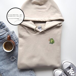 May include: A beige hoodie with a drawstring hood and a small embroidered green frog holding a coffee cup. The hoodie is paired with blue jeans, a coffee mug, and gray sneakers. The logo "CHAMBER OF EMBROIDERY" is in a circle.