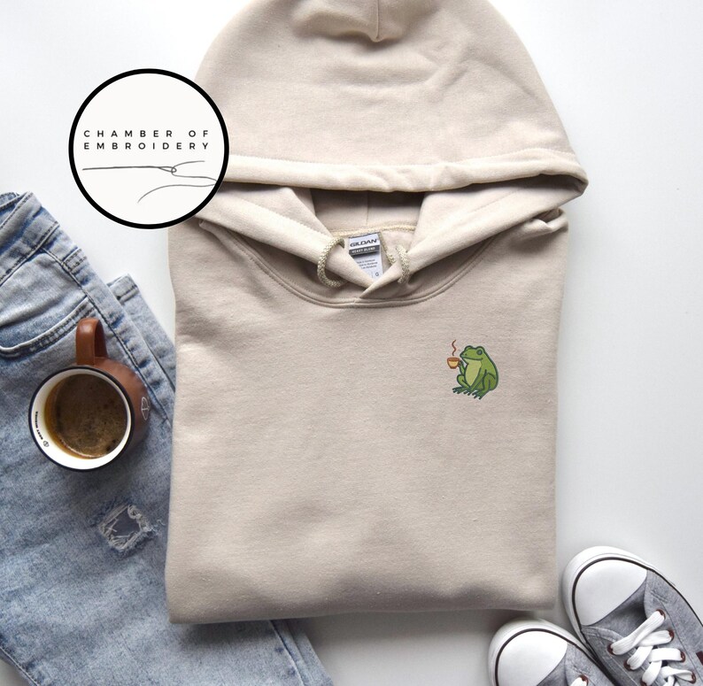 May include: A beige hoodie with a drawstring hood and a small embroidered green frog holding a coffee cup. The hoodie is paired with blue jeans, a coffee mug, and gray sneakers. The logo "CHAMBER OF EMBROIDERY" is in a circle.