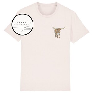 May include: A light pink t-shirt with a brown and white longhorn steer embroidered on the front. The shirt also has a black circle with the text "Chamber of Embroidery" in white.