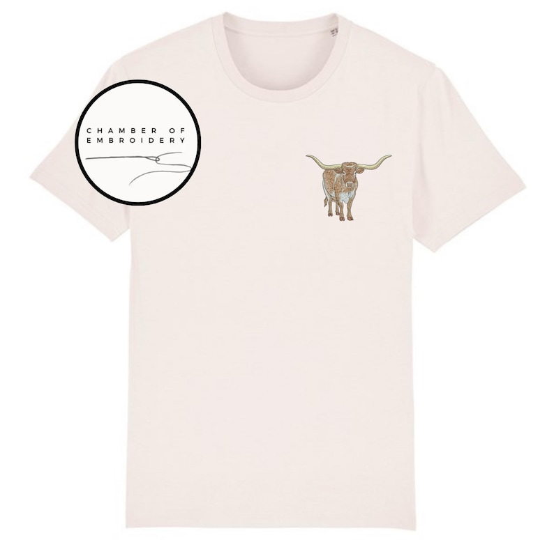 May include: A light pink t-shirt with a brown and white longhorn steer embroidered on the front. The shirt also has a black circle with the text "Chamber of Embroidery" in white.