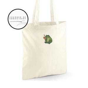 May include: Cream-colored tote bag with a green frog embroidery holding a small brown cup. The bag has two handles and a circular logo that says "CHAMBER OF EMBROIDERY". The frog is detailed with green, brown, and yellow thread.