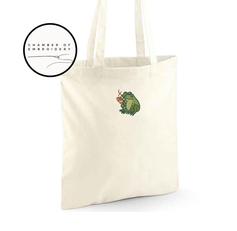 May include: Cream-colored tote bag with a green frog embroidery holding a small brown cup. The bag has two handles and a circular logo that says "CHAMBER OF EMBROIDERY". The frog is detailed with green, brown, and yellow thread.