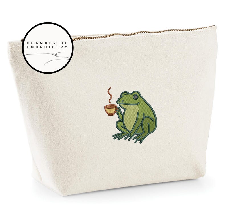 May include: Cream-colored canvas cosmetic bag with a gold zipper. The bag features an embroidered green frog holding a cup of coffee. The bag has the text "CHAMBER OF EMBROIDERY" in a circular logo.
