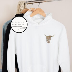 May include: White hoodie with a brown and white longhorn steer embroidered on the front. The hoodie has a drawstring hood and a kangaroo pocket. The text "Chamber of Embroidery" is on a black circle in the top left corner.