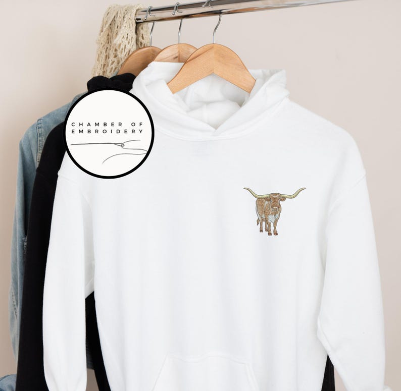 May include: White hoodie with a brown and white longhorn steer embroidered on the front. The hoodie has a drawstring hood and a kangaroo pocket. The text "Chamber of Embroidery" is on a black circle in the top left corner.