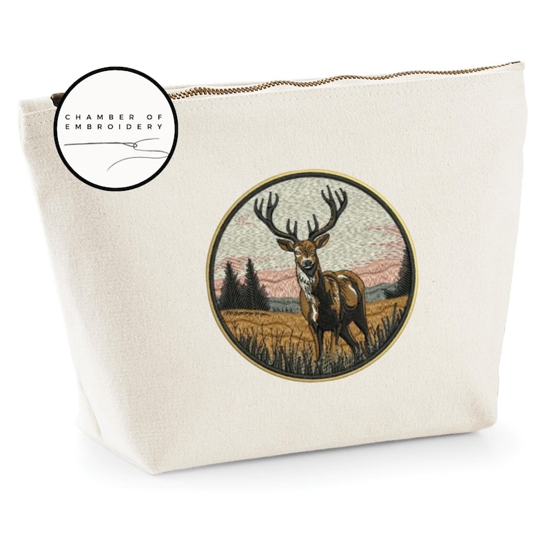 May include: A cream-colored canvas pouch featuring a circular embroidered patch of a stag in a field with trees and a sunset sky. The patch is bordered in gold. A circular logo in the top left corner reads CHAMBER OF EMBROIDERY.
