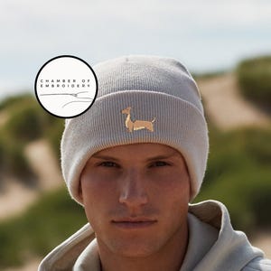 May include: A light gray beanie with a dachshund dog embroidered on the front. The beanie has a ribbed texture and is made of a soft material. The image also includes a circular logo with the text "CHAMBER OF EMBROIDERY".