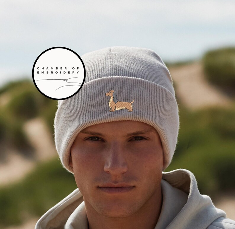 May include: A light gray beanie with a dachshund dog embroidered on the front. The beanie has a ribbed texture and is made of a soft material. The image also includes a circular logo with the text "CHAMBER OF EMBROIDERY".