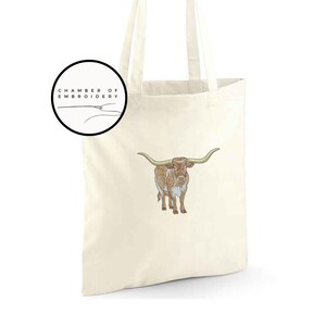 May include: A white tote bag with a brown and white longhorn steer embroidered on the front. The bag has black handles and the text "Chamber of Embroidery" is printed on a black circle in the top left corner.