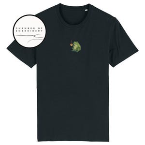 May include: Black t-shirt featuring a small embroidered design of a green frog holding a cup of coffee. The shirt has short sleeves and a crew neck. The logo "CHAMBER OF EMBROIDERY" is in a circle on the left side of the shirt.