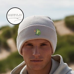 May include: A light gray beanie with a small embroidered frog design holding a cup. The beanie is made of a ribbed knit material. The logo "CHAMBER OF EMBROIDERY" is in a circle in the upper left corner.