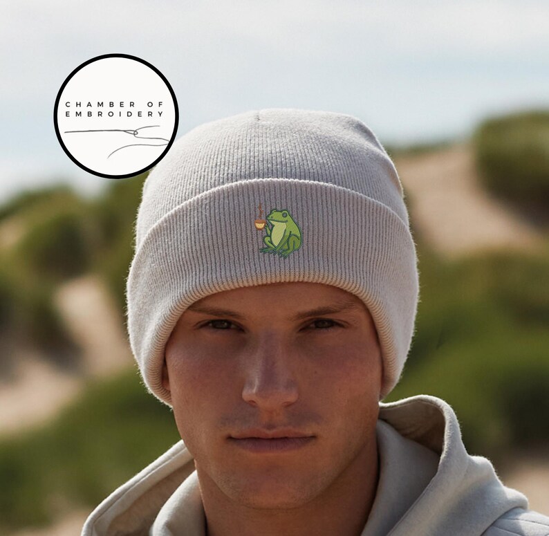 May include: A light gray beanie with a small embroidered frog design holding a cup. The beanie is made of a ribbed knit material. The logo "CHAMBER OF EMBROIDERY" is in a circle in the upper left corner.