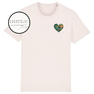 Może przedstawiać: Jasnor&oacute;żowy t-shirt z kr&oacute;tkim rękawem z małym haftem w kształcie serca na lewej piersi, przedstawiającym g&oacute;rski krajobraz z dużą falą i słońcem. W lewym g&oacute;rnym rogu znajduje się okrągłe logo z napisem CHAMBER OF EMBROIDERY.