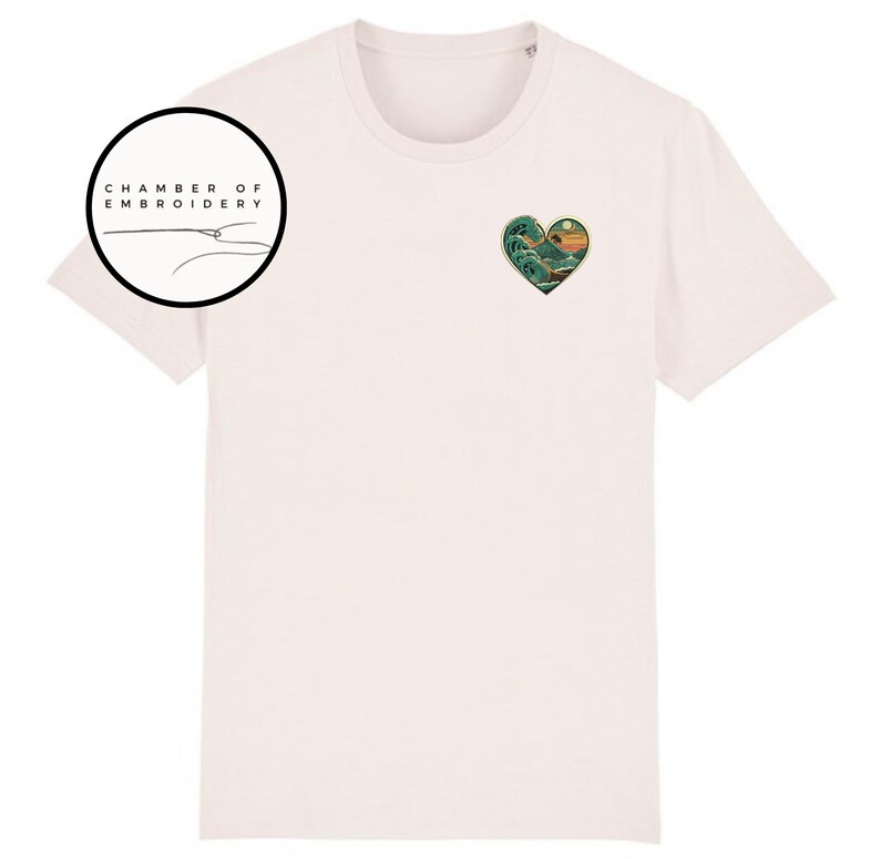 Może przedstawiać: Jasnor&oacute;żowy t-shirt z kr&oacute;tkim rękawem z małym haftem w kształcie serca na lewej piersi, przedstawiającym g&oacute;rski krajobraz z dużą falą i słońcem. W lewym g&oacute;rnym rogu znajduje się okrągłe logo z napisem CHAMBER OF EMBROIDERY.