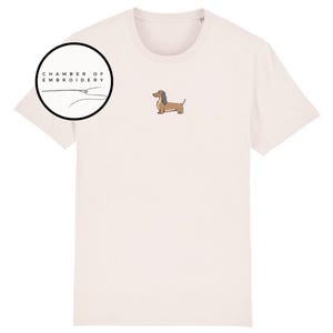 May include: Cream-colored t-shirt featuring an embroidered dachshund dog. The shirt has a crew neck and short sleeves. A circular logo with the text "CHAMBER OF EMBROIDERY" is on the upper left. The dog is brown and tan.