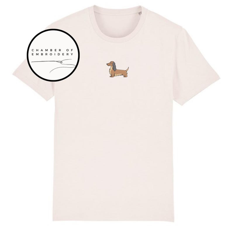 May include: Cream-colored t-shirt featuring an embroidered dachshund dog. The shirt has a crew neck and short sleeves. A circular logo with the text "CHAMBER OF EMBROIDERY" is on the upper left. The dog is brown and tan.