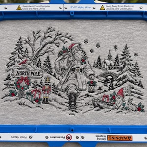May include: A gray fabric panel secured in a blue 8x13 Mighty Hoop embroidery frame featuring a detailed stitched scene of Santa Claus walking through a snowy forest with a nutcracker, gifts, a North Pole sign, and a small gnome.