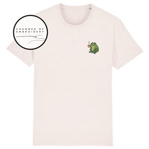 May include: Cream-colored t-shirt featuring a small embroidered design of a green frog holding a cup of coffee. The shirt has a crew neck and short sleeves. The text "CHAMBER OF EMBROIDERY" is in a black circle on the left side.