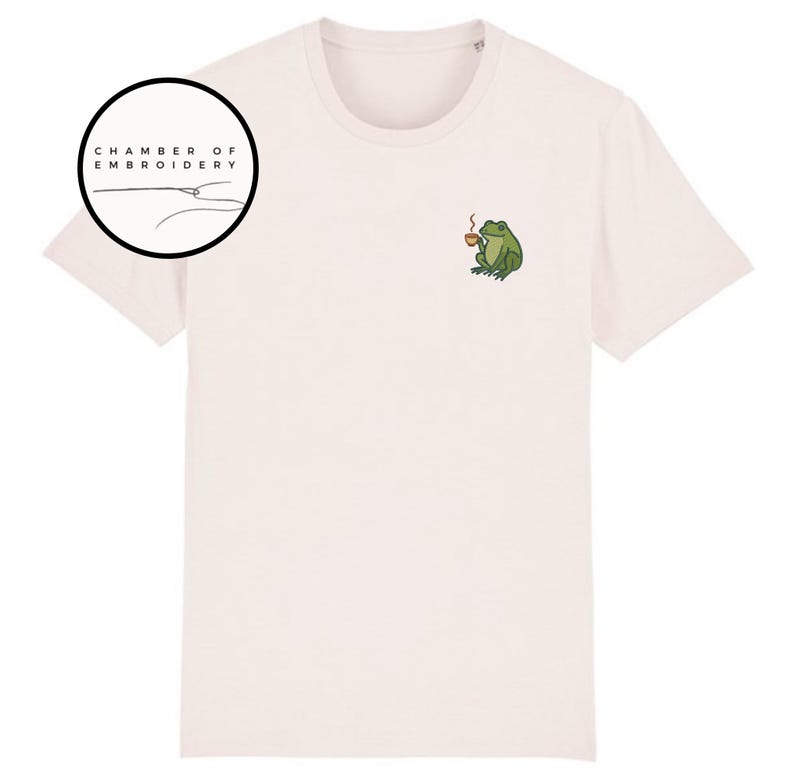 May include: Cream-colored t-shirt featuring a small embroidered design of a green frog holding a cup of coffee. The shirt has a crew neck and short sleeves. The text "CHAMBER OF EMBROIDERY" is in a black circle on the left side.