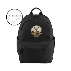 May include: A black backpack featuring a circular embroidered patch on the front. The patch depicts a deer standing in a field with trees and a sunset sky. Above the backpack is a circular logo with the text Chamber of Embroidery.