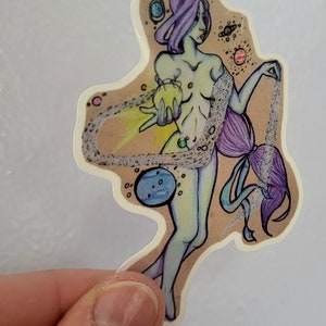 Op de afbeelding: Een sticker met een kleurrijke illustratie van een persoon met lang paars haar die een gloeiende bol vasthoudt. De persoon wordt omringd door een wervelende wolk van sterren en planeten. De sticker is gedrukt op een bruine achtergrond.