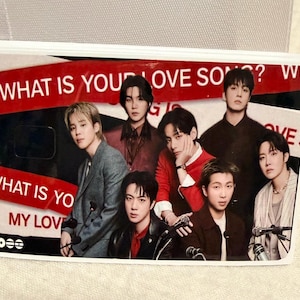 Pu&ograve; includere: Una carta rettangolare con un gruppo di sette persone. La carta ha un testo rosso e bianco che recita "WHAT IS YOUR LOVE SONG?" e "WHAT IS YOUR MY LOVE". Le persone indossano vari abiti.