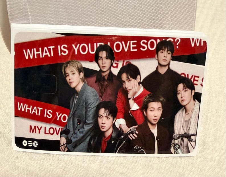 Pu&ograve; includere: Una carta rettangolare con un gruppo di sette persone. La carta ha un testo rosso e bianco che recita "WHAT IS YOUR LOVE SONG?" e "WHAT IS YOUR MY LOVE". Le persone indossano vari abiti.