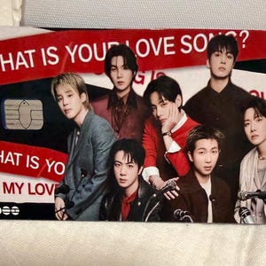 Pu&ograve; includere: Una carta rettangolare con un gruppo di sette persone. La carta ha un design rosso e nero con il testo "WHAT IS YOUR LOVE SONG?" e "WHAT IS YOUR MY LOVE". La carta ha anche un chip.