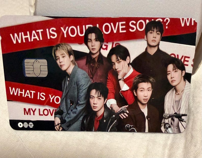 Pu&ograve; includere: Una carta rettangolare con un gruppo di sette persone. La carta ha un design rosso e nero con il testo "WHAT IS YOUR LOVE SONG?" e "WHAT IS YOUR MY LOVE". La carta ha anche un chip.