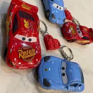 May include: Four toy car keychains featuring characters from the movie Cars. Two red cars with Rust-eze branding and the number 95, and two light blue sports cars. Each car is attached to a metal keyring with a small red plastic brick connector.