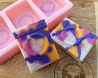 Op de afbeelding: Twee kleurrijke, handgemaakte zeepjes in de vorm van wasmachines. De zeepjes zijn versierd met swirls van paars, geel, roze en wit. De zeepjes liggen op een houten oppervlak.