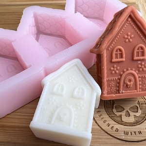 May include: Two gingerbread house moulds, one pink and one white, with a brown gingerbread house in the centre. The moulds are made of silicone and have a detailed design. The gingerbread house is decorated with white icing and red sweets.