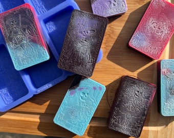 Puede incluir: Seis jabones con forma de cartas de tarot con diferentes diseños y colores. Los jabones están hechos en un molde de silicona y presentan imágenes de esqueletos, cráneos y otras imágenes espeluznantes. Los jabones están etiquetados con los nombres de las cartas de tarot: "Muerte", "El Mago", "Los Amantes", "El Loco", "Juicio" y "El Ahorcado".