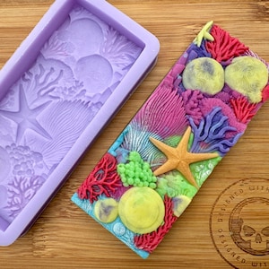 May include: A purple silicone mold with a starfish and coral design. The mold is next to a finished soap bar with a similar design, featuring coral, starfish, and seashells in shades of pink, purple, green, and yellow.