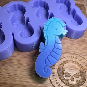May include: A silicone mold for making seahorse-shaped soap or resin. The mold is purple and has five seahorse cavities. A blue and green seahorse is sitting on top of the mold.